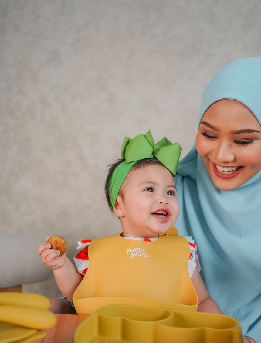 Baby laughing in her Plate and Play bib, a bite of food in hand, her mum smiling beside her